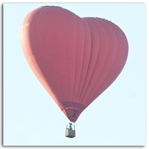 Ein einzelner Heißluftballon in Herzform in Nahaufnahme vor hellem Himmel. Der Ballon ist vollständig rot und trägt in weißen Buchstaben die Aufschrift „LOVE“.