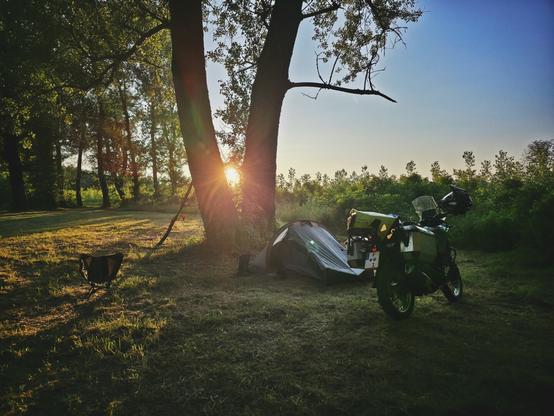 A low-angle, landscape-oriented photo shows a campsite scene at sunset. In the foreground, a grey tent is pitched to the right of two large trees with sunlight peaking through their trunks. To the right of the trees and the tent, a motorcycle with gear strapped to its back is parked. A small, dark portable chair is visible to the far left. The ground is covered in green and yellow grass. In the background, sunlight illuminates the trees and a hazy sky, with a line of smaller trees or bushes visible further back.