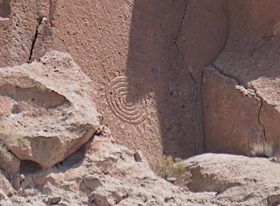 A petroglyph of six concentric circles with rays radiating out from the outermost circle, carved in a vertical, very flat wall of soft volcanic tuff. A triangular shadow is coming from above and right, covering part of the circle, with the tip of the triangle ending up exactly on the center of the circles.