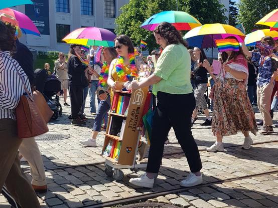 Real photograph of two librarians, I presume, with a trolley marked "Biblioteket for alle" (translates to "The library for all"). Many rainbow-coloured umbrellas (or parasols, the sun is bright!). Real photographer: me.