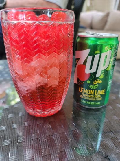 This is a close-up, eye-level shot of a red beverage, Moonshine Cherry, in a textured glass, positioned next to a small can of 7 UP. Both items are sitting on a dark, reflective, possibly glass table.

The glass on the left is tall and clear, with a textured, chevron-like pattern embossed on its exterior, creating a ribbed effect. It's filled with a vibrant, translucent red liquid,  a cherry-flavored moonshine drink, given the several bright red maraschino cherries floating on top. The drink also appears to contain crushed ice, contributing to its frosty appearance.

To the right of the glass is a small, green can of 7 UP Lemon Lime flavored soda. The can features the iconic 7 UP logo with a red circle and green lettering. It has a slightly dulled, metallic sheen.

The background is softly blurred, showing hints of what appears to be an outdoor setting, possibly a patio, with some light-colored upholstery or furniture visible. The overall lighting suggests it's daytime.