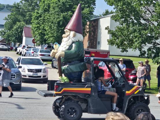 One of the Gnome Peterson statues with a red hat, white beard and green tunic, holding a blue and white bird,  riding the back of a small four wheeled vehicle.