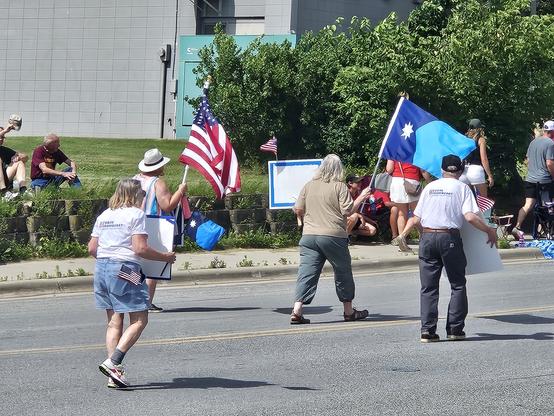 Minnesota and American flag and 4 people with 3 signs honoring the DFL  State Rep Melissa Hortman, her husband and dog who were murdered by a Republican