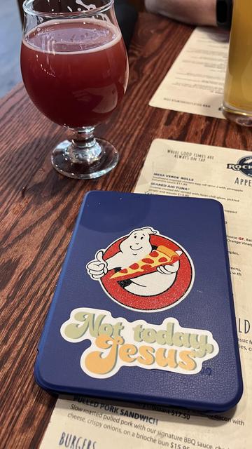 The image shows a close-up of a table with various items. In the foreground, there is a blue case with a cartoon character holding a slice of pizza, accompanied by the text "Not today Jesus." The case is placed on a wooden table. To the left, there is a glass containing a reddish beverage, possibly a craft beer, with a frothy head. In the background, a menu is partially visible, featuring items like "Mesa Verde Rolls" and "Seared Ahi Tuna." Another glass, possibly containing a yellowish beverage, is also visible. The overall setting suggests a casual dining environment.

Provided by @altbot, generated privately and locally using Ovis2-8B