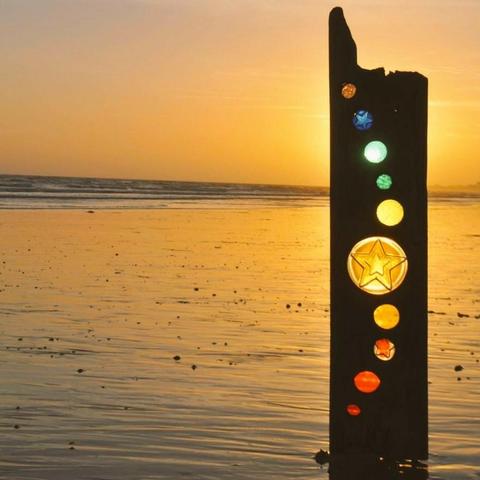 Rectangular driftwood vertical post inlaid with circles containing coloured stained glass, one with a central star motif,, the sculpture is sited on a wet sandy golden shoreline at dawn