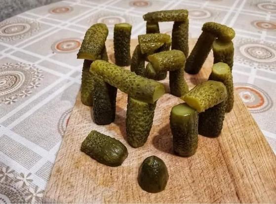 A wooden cutting board displays creatively arranged pickles, resembling stones in a mini Stonehenge structure. The background features a patterned tablecloth.

Ein hölzernes Schneidebrett zeigt kreativ angeordnete Gurken, die Steinen in einer Mini-Stonehenge-Struktur ähneln. Der Hintergrund zeigt eine gemusterte Tischdecke.