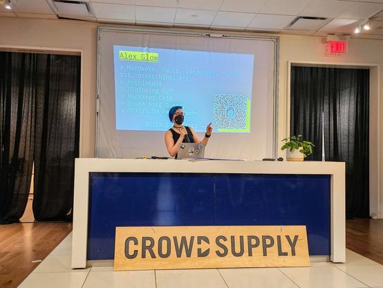 Merlin in front of a slide listing her socials, with a Crowd Supply sign in front of that