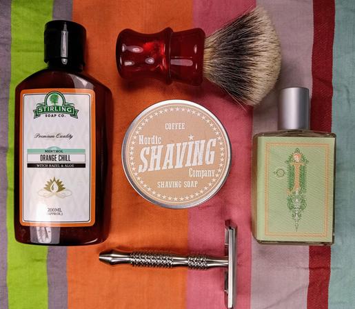 (Le Chat Mistral description)

The image showcases a collection of grooming items arranged on a striped background. At the top left, there's a brown bottle of aftershave with a white label reading "Stirling Soap Co. Orange Chill." To the right of the bottle, there's a red shaving brush with a wooden handle. Below the brush, in the centre, is a round tin labelled "Nordic Shaving Company Shaving Soap." To the right of the tin, there's a small bottle of cologne with a vintage design. At the bottom, there's a silver safety razor with a textured handle. The items are neatly arranged, creating a visually appealing setup for a traditional shaving routine.