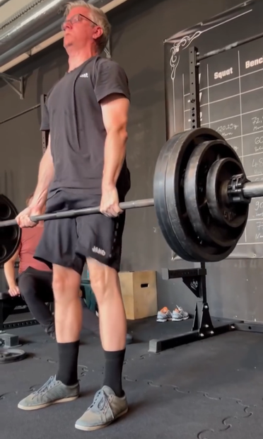 A middle-aged man in a black adidas t-shirt and shorts is performing a deadlift in a gym. He is gripping a barbell loaded with several large weight plates, pulling it up from the floor. His head is tilted slightly back and his red face and intense facial expression shows his effort. In the background, there's a squat rack with a whiteboard showing numbers for "Squat" and "Bench" in a powerlifting gym.