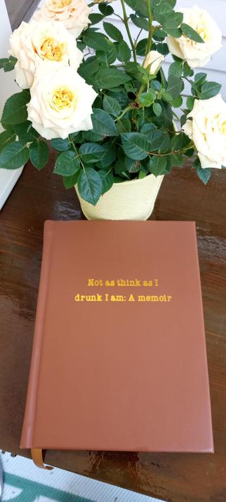 A tan leather book, A4 size. A title is embossed on the cover with gold leaf: "Not as think as I drunk I am: A memoir." The book is on a wood bar with a small yellow rose plant in the background.