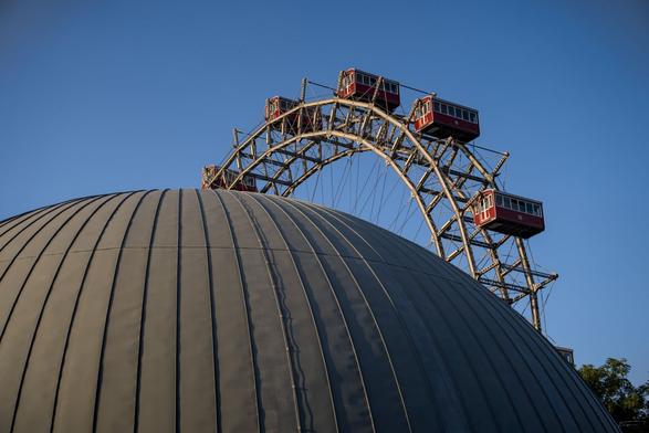 Das Bild zeigt das Wiener Riesenrad im Prater, eines der bekanntesten Wahrzeichen der Stadt. Im Vordergrund befindet sich die markante, gerippte Kuppel des Planetariums der Stadt Wien, die einen Teil des Riesenrads verdeckt. Die nostalgischen roten Gondeln des Riesenrads heben sich schön vom klaren blauen Himmel ab und bilden einen spannenden Kontrast zur modernen Architektur des Planetariums. Die Aufnahme fängt auf eindrucksvolle Weise den Übergang zwischen Wissenschaft, Technik und Wiener Geschichte ein.