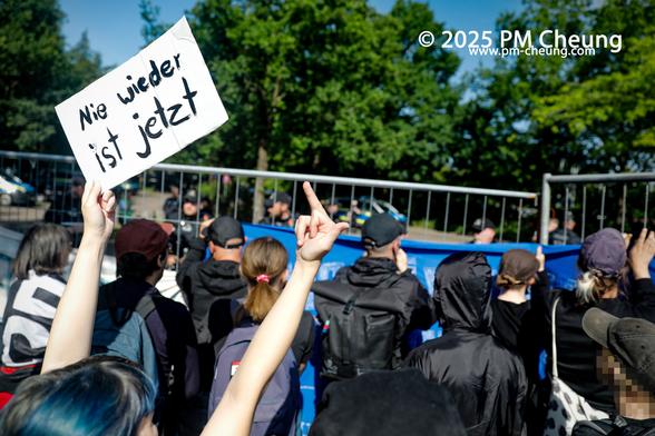 Am Bürgerhaus standen sich die Gegendemonstrant*innen und die Delegierten der AfD gegenüber. Eine Demonstrant*in reckt ein Schild mit der Botschaft "Nie wieder ist jetzt" in die Luft. Gleichzeitig zeigt sie auch ihren Mittelfinger in Richtung der AfD.