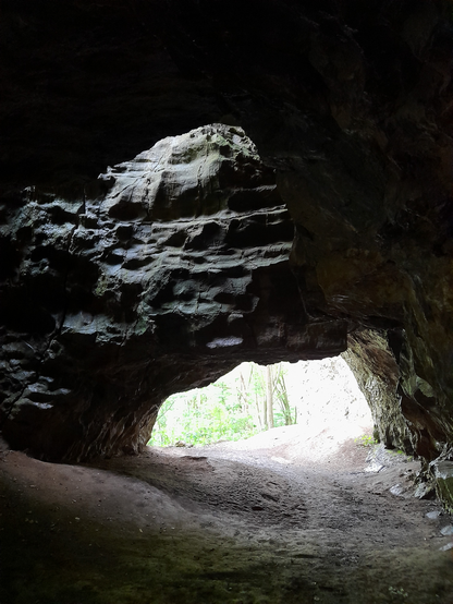 A view from inside a cave tunnel. It is dark, but some light is coming from a hole in the cave roof and further away and lower the bright sun light barely makes some trees visible.