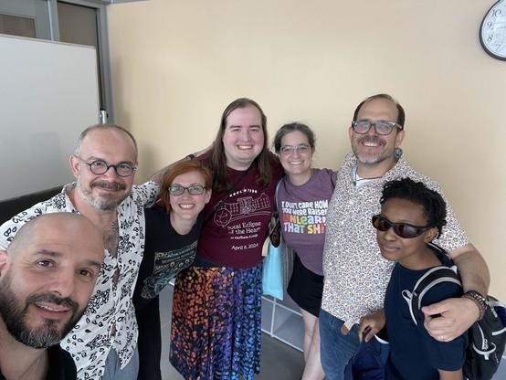 7 people gather in front of the camera. A bald bearded man is clearly holding the camera for the selfie. Behind him a paler man with a grey beard and mustache and round glasses smiles. Below his arm peeks a grinning redhead with rectangular glasses. Next is a pale brunette with a maroon shirt and rainbow speckled skirt. Next is a bespectacled woman with salt and pepper hair and a purple shirt that says “I don’t care how you were raised. Unlearn that shit.” Then a taller, more tanned, bearded man with glasses and a necklace has his arm around a shorter person with darker skin, locs, and sun glasses.