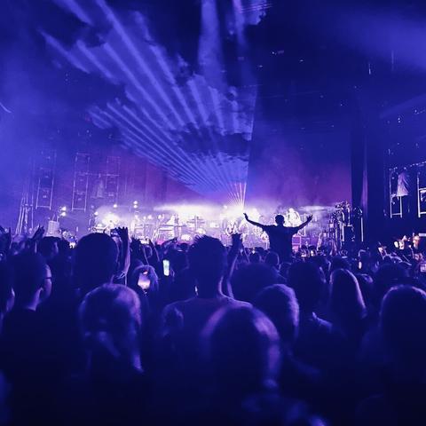 shot from a crowd viewing a stage. you can barely make out the band. laser lights appearing above the crowd. a person is on someone’s shoulders with their arms in the air. everything looks blue and purple in colour.