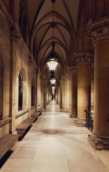 Cityhall of Vienna.

A long, dimly lit corridor with arched ceilings and stone walls, featuring lanterns hanging from the ceiling and benches along the sides. Columns line the walkway, creating a symmetrical perspective.