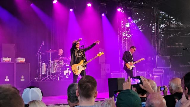 Glenn Hughes (Gesang, Bass), Joe Bonamassa (Gitarre) und Jason Bonham (Schlagzeug) live auf der Bühne beim Konzert von Black Country Communion am Tanzbrunnen Köln. Die Bühne ist in violettes Licht getaucht, das Publikum feiert, fotografiert und filmt. Energiegeladene Rock-Atmosphäre unter freiem Himmel.