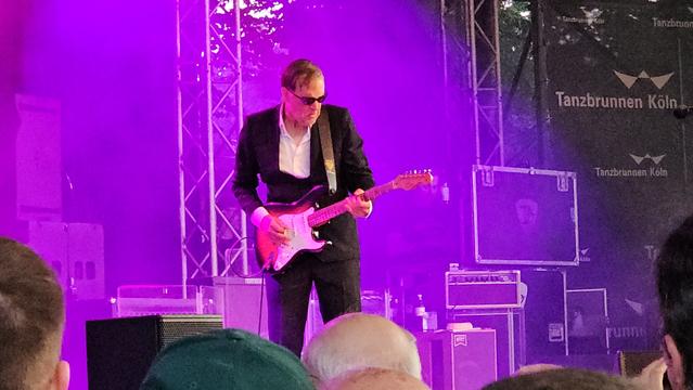 Joe Bonamassa bei einem Auftritt von Black Country Communion am Tanzbrunnen Köln – im Anzug, mit Sonnenbrille und Stratocaster in violettes Bühnenlicht getaucht. Gitarrensolo-Intensität trifft auf Open-Air-Stimmung.