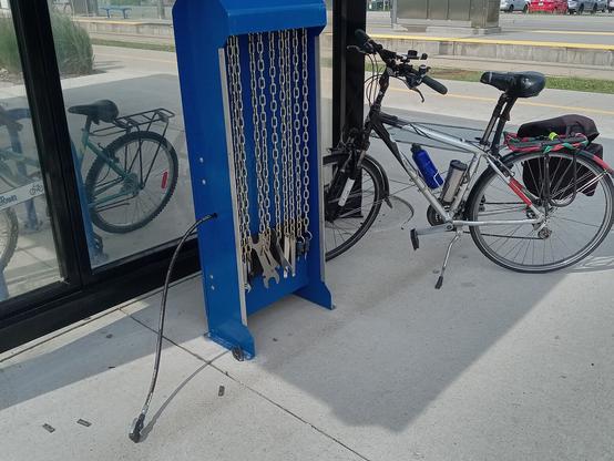 Bike repair station at Willis Way LRT stop. The inflator hose is on the pavement, but the nozzle still looks intact. Bob's bike is in the background, and a bike is parked in the shelter.