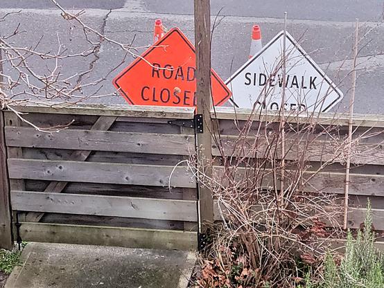This image shows the edge of our front garden, with the fence and gate. On the sidewalk side of the gate are two construction signs, both facing the house. The first reads, "Road Closed". The second reads, "Sidewalk Closed". We tried really hard not to take it personally.