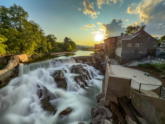 The sun sets behind a large waterfall from a dam next to a mill