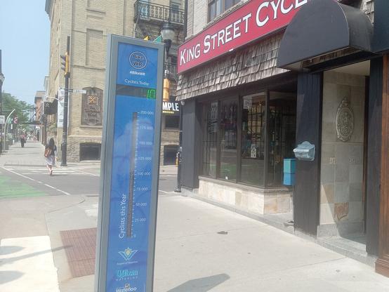 A blue, upright bike counter in Uptown Waterloo, showing "10" (but the display was actually 110, probably lost a digit because of shutter speed unsynced with the LED). Logos for #WaterlooRegion are visible at the bottom of the post.

The protected bike lane recedes to the left, the King Street Cycles LBS is on the right.