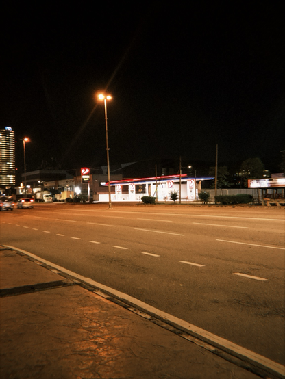The image depicts a nighttime urban scene with a wide, multi-lane road in the foreground. The road is illuminated by streetlights, casting a warm glow on the asphalt. On the right side of the road, there is a brightly lit gas station with a distinctive red and white color scheme and the logo "D2" prominently displayed. The gas station has a canopy with red and blue lighting, and several fuel pumps are visible. In the background, there are other buildings, including a tall, illuminated skyscraper on the left side of the image. The sky is dark, indicating it is nighttime. The overall atmosphere is quiet, with no visible pedestrians or heavy traffic.