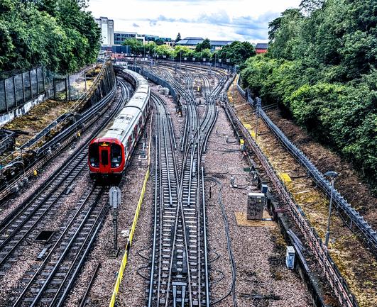 A metropolitan line tube train part of tfl coming up a gentle slope to go under a bridge. Having just left Uxbridge station. The tube lines look very black and shiny from the recent rain to the right of the picture. Are the sidings used by the tube trains overnight.