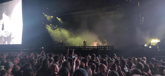 Nine Inch Nails op de South Stage op Graspop 2025. Links zijn er op een enorm scherm zwart-wit beelden van te zien. Het podium wordt geelgroen verlicht. Op de voorgrond publiek, sommigen met hun hand in de lucht.