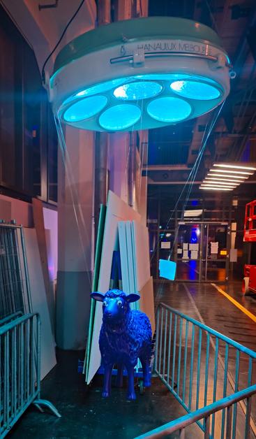 Ein blaues Schaf schwebt circa 1m über dem Boden und wird von einer großen Lampe angeleuchtet. Im Hintergrund ist ein Laufweg mit Leitstreifen und ein Rollwagen mit Materialen zu sehen.