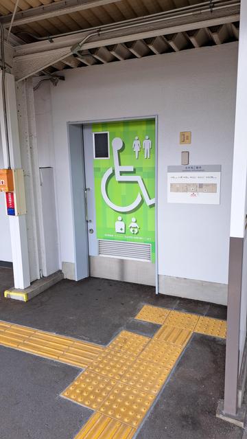 A picture of the center bathroom door, which is green with a large handicapped symbol and various smaller symbols indicating that it's gender-neutral and suitable for changing babies and dealing with special medical needs. Visible on the ground are large yellow tactile paving blocks that lead right up to the guidemap.