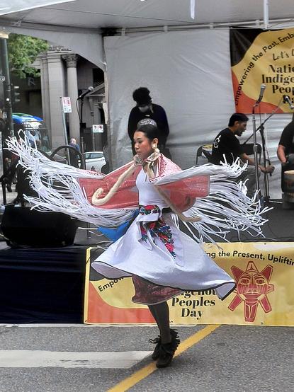 A shawl dancer twirls to a drum beat as white ribbons follow in motion.
