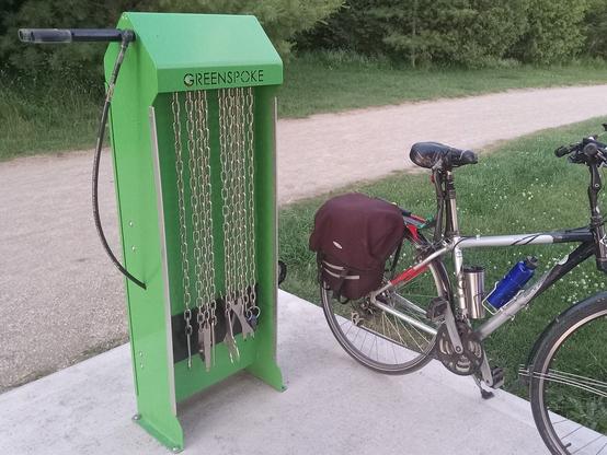 A green bike repair station on a concrete pad. Behind is a crushed gravel trail (the Health Valley / Trans Canada Trail). There is lots of greenery on the other side of the trail, and some short grass to the right behind Bob's bike.
