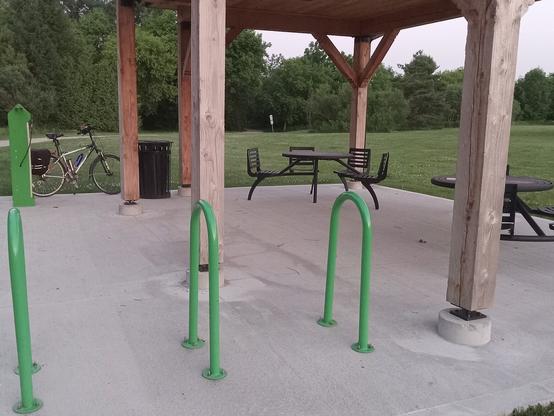 Picnic shelter at the end of the Health Valley / Trans Canada Trail in St. Jacobs. There are three green bike racks ("staples") at the front of a concrete pad which is maybe 10x10 metres. At the back right is a green bike repair station with Bob's bike behind it.  Also at the back there's a black garbage can, and two circular metal tables with attached chairs. There are wooden posts regularly spaced on the concrete pad holding up the shelter roof. In the background is a field with closely cropped grass, and even further back is a thick forest of trees. 

Everything looks very new and antiseptic.