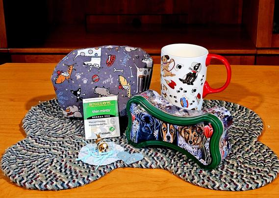 Tea tableau on a braided mat shaped like a dog biscuit. There are a cozy and a mug with cartoon dog designs. A bone shaped tin with portraits of dogs, a tiny figurine of a dog on a rug naughtily shredding paper. The tea is Thin Mints Girl Scout Cookie black tea from Bigelow.