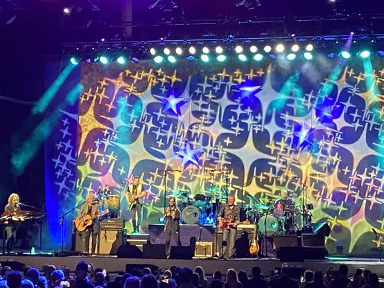 Ringo Starr and His All Starr Band performing in front of another 1960s psychedelic backdrop, with a kaleidoscope esque pattern.