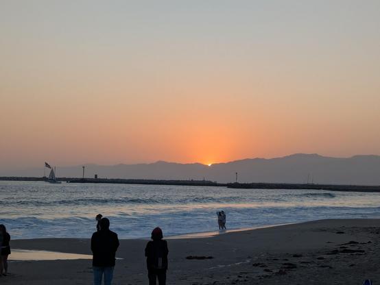 The sun sets over the Pacific ocean as waves roll in. The water is blue. The sky is orange turning to greenish blue at the top of the frame. Several silhouetted people stand in the middle ground.