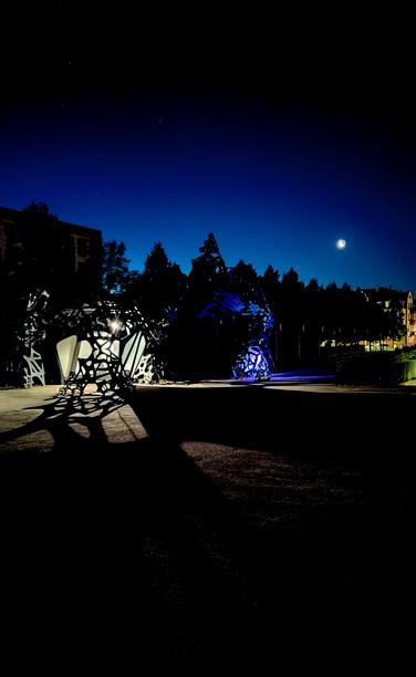 Foto in den Morgenstunden. Licht strahlt durch aus Blech geformte Strukturen und wirft Schatten auf dem leeren Vorplatz des ZKM. Im Hintergrund ist der Mond zu sehen.