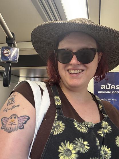 Photo of me smiling with sunglasses and a sun hat on. I am in a train, with handrails visible behind me.