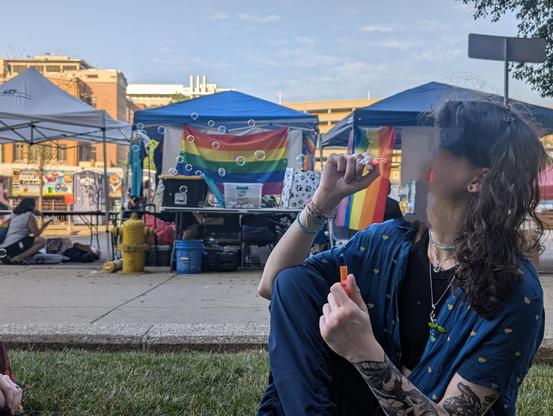 A picture of this enby sitting in a patch of grass near a sidewalk. They are blowing bubbles across the frame from right to left. Holding the bubble blow stick with their right hand, they're resting their right inner elbow against the outer right knee to control the angle with their hips while keeping the hand and frame stable. Colorful beads and metal bracelets decorate their right forearm while their left hand is holding the bubble solution in an orange vial. Her left forearm is covered in nature tattoos, the leaves at the wrist act as a nest for a potion bottle sealed with moss. You can make out part of the owl with portals in it. You can also see the toad on the toadstool by the bicep. She's wearing the blue slouch pants, black crop, and blue collared short sleeve shirt with rainbow hearts. There is a sprout on the shirt now, near the salamander necklace. They are also wearing a non-binary pendant and multicolored bead choker. Their messy coils/waves cover most of the rest of the side, but you can make out that they are wearing their green plugs. 

In the background you see a few booths for the  Pride event, and farther back is the food truck zone.