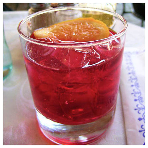 he image features a close-up of a glass containing a red liquid, likely a cocktail, with ice cubes floating inside. The glass is placed on a white tablecloth with a subtle blue pattern. A slice of orange is garnishing the drink, resting on the rim of the glass. The background is slightly blurred, suggesting a dining setting with other items, such as a bottle, visible but out of focus. The overall composition highlights the vibrant red color of the drink, the transparency of the glass, and the texture of the ice and orange slice.

Provided by @altbot, generated privately and locally using Ovis2-8B
