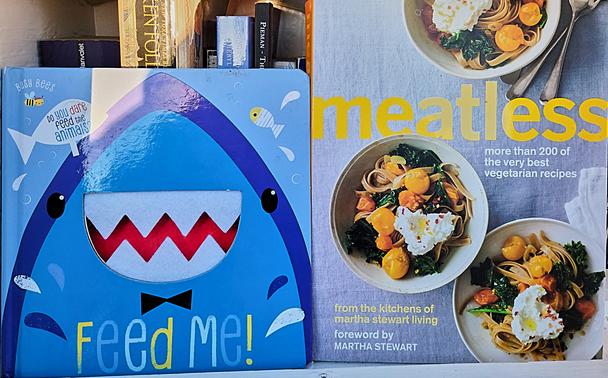 This image shows two books that were found side by side in a local Little Library. The book on the left reads "FEED ME!" and has an image of a playful shark on the cover. The book to the right has the title "Meatless" and contains over 200 vegetarian recipes. Together they bring to mind the scene from the movie "Finding Nemo".