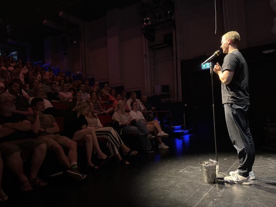 Julius Althoetmar: Ein Poetry Slammer mit kurzem blondem Haar, schwarzem T-Shirt und dunkler Hose steht auf der Bühne. Er spricht ins Mikrofon und gestikuliert leicht. Alle Blicke sind auf den Slammer gerichtet. 
Die Brechtbühne des Staatstheater Augsburg ist beim GRAND SLAM – Der Poetry Slam in Augsburg voll besetzt.