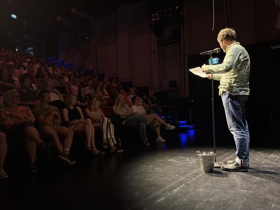 Christian Kreis: Ein Slammer mit leicht zerzausten Haaren, gemustertem Hemd und Jeans steht auf der Bühne. Er liest von Blättern und spricht ins Mikrofon. Das Publikum sitzt eng und hört konzentriert zu beim GRAND SLAM – Der Poetry Slam in Augsburg in der brechtbühne des Staatstheaters.