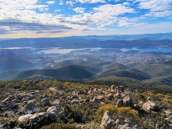 Scattered clouds over a view from the top of mountain over hills and a river, rocks and shrubs in the foreground.