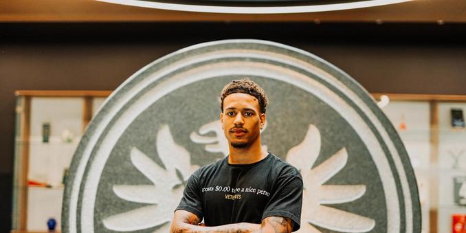 Ein junger Mann mit kurzen, welligen Haaren und einem ernsten Gesichtsausdruck steht vor einem großen, runden Emblem mit einem stilisierten Vogel, der nach oben fliegt. Er trägt ein schwarzes T-Shirt mit der Aufschrift "costs $0.00 to be a nice person VETERANS". Seine Arme sind verschränkt, und er hat Tattoos auf seinen Armen. Im Hintergrund sind Regale mit verschiedenen Gegenständen zu sehen, die in einer modernen, gut beleuchteten Umgebung platziert sind.