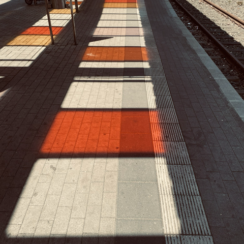 Blick auf einen Bahnsteig: Schatten des Glasdaches, in dem einige farbige Glasplatten eingesetzt sind (warme Farben, vor allem rot und orange)