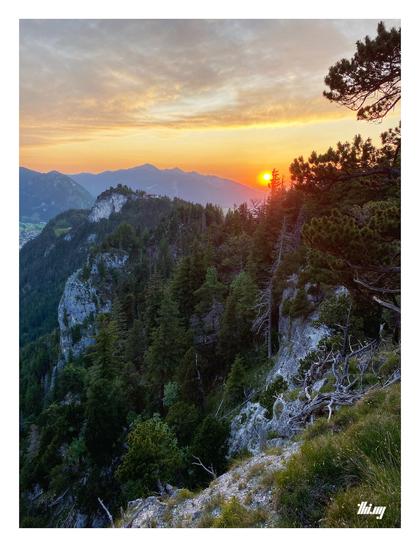 Photo of an alpine ridge line with steep rocky cliffs and lush pine forest. The sun is almost dipping behind the mountains in the back and the sky is illuminated in golden glow.