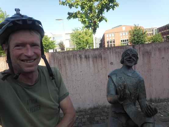 Selfies eines Radlers mit Helm und olivfarbenem Shirt, sitzend neben einer Bronzeskulptur.