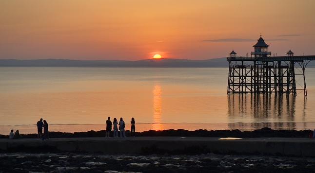 A group of people stand along a sea wall in Clevedon, watching the sun set over calm water. The glowing orange sun dips just below the horizon, casting a warm reflection across the sea. To the right, Clevedon Pier—an elegant Victorian-style structure—extends over the water, its silhouette outlined against the golden sky. The scene is tranquil, with warm amber and orange tones creating a peaceful atmosphere.